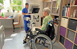 Durch den „Teledoc-Rollständer“ können Pflegefach- und Pflegehilfskräfte schnell und unkompliziert per Bildschirm miteinander kommunizieren. Foto: Vereinigte Hospitien Eine Pflegerin in blauer Berufskleidung kümmert sich um eine Seniorin im Rollstuhl und benutzt dabei einen Bildschirm, der auf einer Rollvorrichtung montiert ist.
