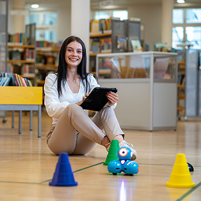 Junge Frau steuert in der Bibliothek mit einem Tablet einen kleinen Roboter