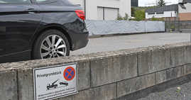 Eine Tafel auf einer niedrigen Mauer weist auf das Parkverbot auf einem Privatparkplatz hin, der im Hintergrund ebenso wie das Heck eines Autos zu sehen ist.